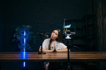 Photo for Funny female blogger sitting at the table at home illuminated by professional lighting, speaking into a microphone and looking at the camera, recording videos on YouTube - Royalty Free Image