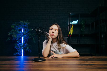 Photo for Attractive female blogger sitting with thoughtful face near microphone in studio and looking away. - Royalty Free Image