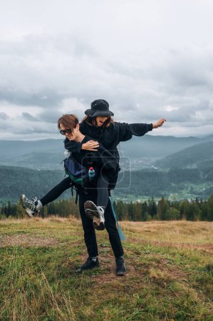 Photo for A cheerful couple of young tourists in casual clothes are having fun on the top of the mountain, the man lifted the woman on his back and carries on the background of beautiful mountain views. - Royalty Free Image