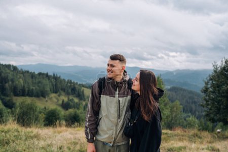 Photo for A positive couple, man and woman, are standing in the mountains on the meadow against the background of beautiful Carpathian views and looking away with a smile on their faces. - Royalty Free Image