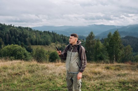 Photo for A handsome young male tourist in casual clothes stands in the mountains with a smartphone in his hands and looks at the route where to go next. - Royalty Free Image