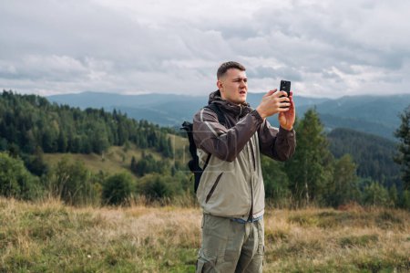 Photo for Sad young man tourist with smartphone in hands stands in the mountains during a hike and catches a cellular network. - Royalty Free Image