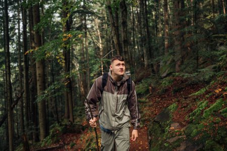 Photo for A handsome male tourist in a jacket and with a backpack on a mountain hike climbs through the forest on a path with a stick in his hands. - Royalty Free Image
