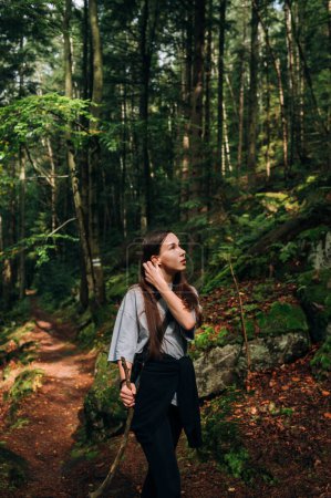Photo for Beautiful woman in casual clothes stands in the middle of the forest in the mountains and rests on a hike in the sun. - Royalty Free Image
