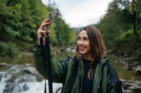 Photo for Happy beautiful woman tourist in the mountains on the background of the river takes a selfie on the smartphone camera with a smile on her face, looks away and laughs. - Royalty Free Image