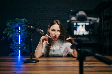 Photo for Backstage photo of a female blogger sitting at a table. in the studio with a microphone and recording a podcast video with an emotional funny face, looking at the camera. - Royalty Free Image
