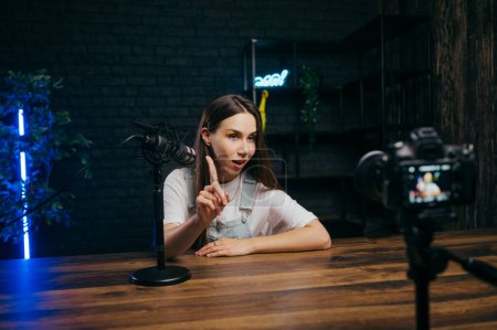 Photo for Attractive emotional female blogger recording video blogs at a table in a studio, speaking into a microphone and pointing a finger at the camera. Backstage photo - Royalty Free Image