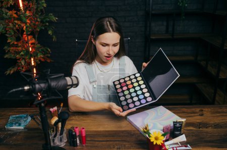 Photo for Surprised female blogger with eyeshadow palette in hands shows makeup to camera and speaks into microphone while recording videos. - Royalty Free Image