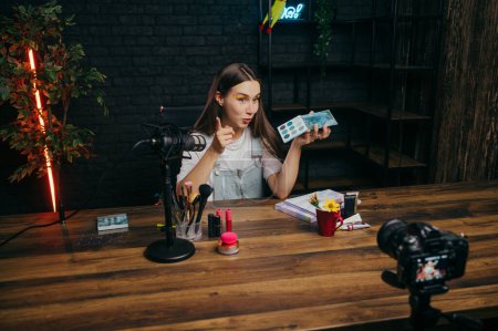 Téléchargez les photos : Dans les coulisses de la blogueuse beauté assise en studio à table avec des cosmétiques et parlant de palette de fards à paupières à la caméra dans le microphone - en image libre de droit