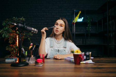 Photo for Portrait of an attractive beauty blogger woman sitting in the studio at the table with a brush in her hand and filming a make-up tutorial, looking at the camera. - Royalty Free Image