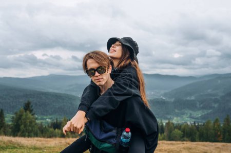Photo for Young beautiful couple man and woman in dark casual clothes having fun in the mountains during a hike, the guy is carrying the girl on his back and looking at the camera. - Royalty Free Image