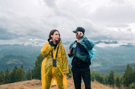 Photo for A young couple of active tourists in casual clothes stand in the mountains wearing raincoats against the background of cloudy views and look in love - Royalty Free Image