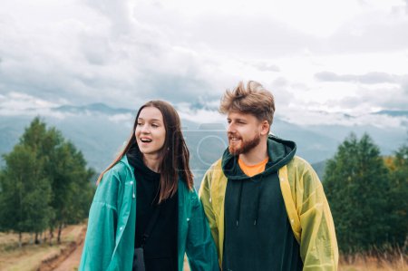 Photo for Photo of a happy couple of tourists on a hike standing on the top of a mountain wearing raincoats and looking away with a smile on their face - Royalty Free Image