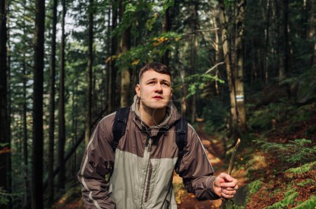 Photo for Handsome young man with a stick in his hands is traveling in the mountains, walking on a path in the forest and looking ahead - Royalty Free Image