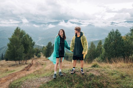 Photo for Young beautiful couple in raincoats holding hands standing in the mountains against the background of beautiful views and looking at beautiful scenery during a hike - Royalty Free Image