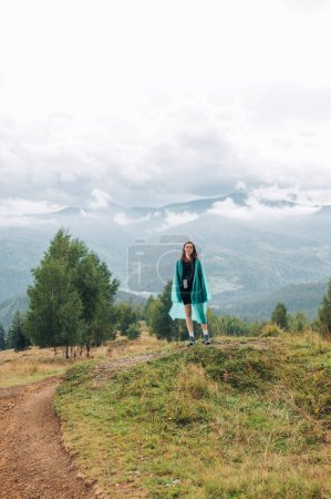 Foto de Foto vertical de una turista en un impermeable de pie en las montañas sobre un fondo de vistas nubladas y mirando a la cámara. Concepto de senderismo - Imagen libre de derechos