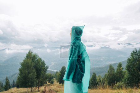 Téléchargez les photos : Une touriste en imperméable se tient dans les montagnes et regarde loin avec un visage sérieux sur fond de vue sur la montagne. - en image libre de droit