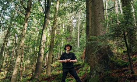 Téléchargez les photos : Jeune randonneur masculin positif en vêtements décontractés debout dans la forêt tout en escaladant les montagnes et en regardant la caméra avec un sourire sur son visage. - en image libre de droit