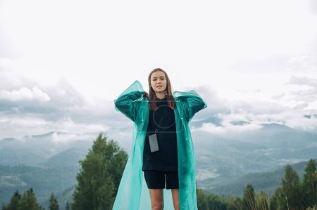 Photo for Portrait of a female tourist in a raincoat stands in the mountains against the background of overcast views and poses for the camera with a serious face. - Royalty Free Image