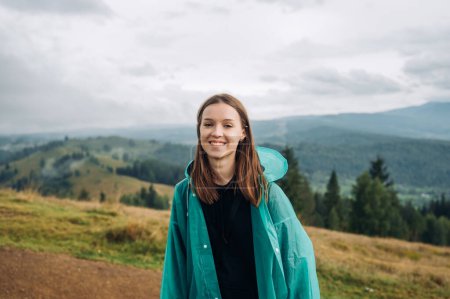 Foto de Retrato de una hermosa turista feliz en una caminata de pie en las montañas en un impermeable sobre un fondo de hermosas vistas nubladas y mirando a la cámara con una sonrisa en su cara. - Imagen libre de derechos