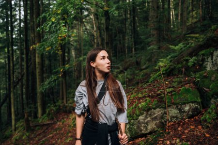 Cute woman hiker in casual clothes stands in the mountain forest on a hike and looks away with a serious face.