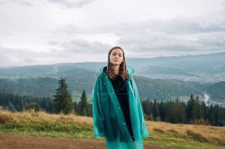 Téléchargez les photos : Attrayant touriste féminine dans un imperméable bleu se tient avec un visage sérieux dans les montagnes sur le fond de belles vues et pose pour la caméra. - en image libre de droit