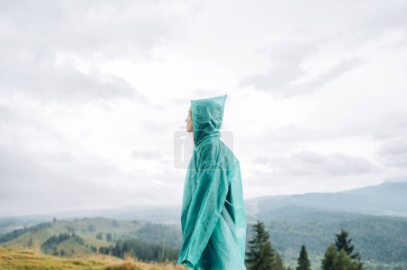 Photo for Woman tourist hiking in the mountains stands in a blue raincoat in gloomy rainy weather and looks away with a serious face. - Royalty Free Image