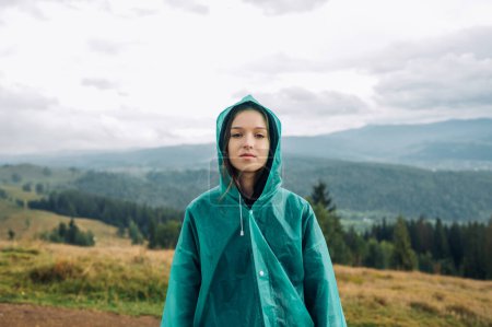 Photo for Female tourist in a blue raincoat stands in the mountains in rainy weather during a hike and looks at the camera with a serious face. - Royalty Free Image