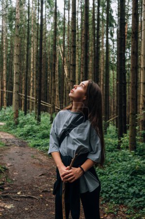 Téléchargez les photos : Une touriste avec un bâton dans les mains se tient dans une forêt de pins et regarde les beaux arbres. Photo verticale - en image libre de droit