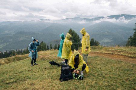 Photo for A group of tourists in raincoats on the top of a mountain stand and prepare for a hike against the background of unreal landscapes with clouds and fog. - Royalty Free Image