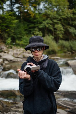Photo for A hipster man with a digital camera in his hands takes a video on a trip against the background of a mountain river, a tourist films his trip to the mountains with a serious face. - Royalty Free Image