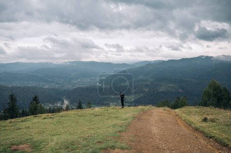 Photo for Photo of a man in dark casual clothes standing in the mountains against a background of beautiful landscapes with mountains and a village. - Royalty Free Image