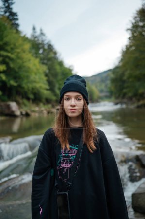 Téléchargez les photos : Portrait d'une belle femme en tenue décontractée posant pour la caméra sur le fond d'une rivière de montagne et une belle vue sur la forêt, regardant la caméra avec un visage sérieux. - en image libre de droit