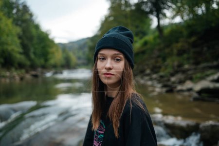Photo for Attractive female tourist in a hat stands on the bank of a fast mountain river and poses for the camera against the background of a beautiful landscape, close-up portrait. - Royalty Free Image