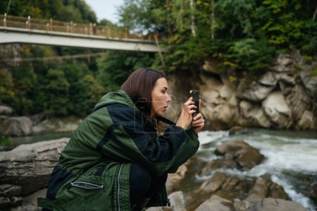 Foto de Turista sentada en una roca cerca de un río de montaña y toma una foto en una cámara de teléfono inteligente con una cara seria - Imagen libre de derechos