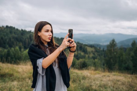 Photo for Attractive female tourist hiking in the mountains stands with a smartphone in her hands and takes a photo on the camera with a serious face and looks away on the background of landscape with mountains - Royalty Free Image