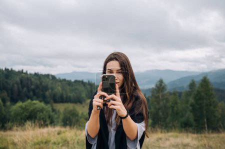 Beautiful woman in casual clothes stands with a smartphone in her hands in the mountains and takes a photo with a serious face.