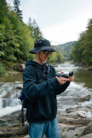 Foto de Foto de un elegante turista masculino en las montañas filmando un video de viaje en una vieja cámara digital sobre el fondo de un río de montaña. - Imagen libre de derechos