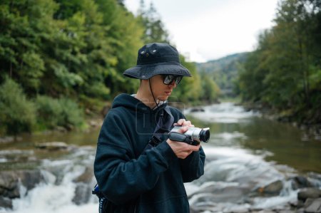 Téléchargez les photos : Élégant gars touriste en panama et vêtements décontractés tourne une vidéo dans les montagnes sur un vieil appareil photo numérique. - en image libre de droit