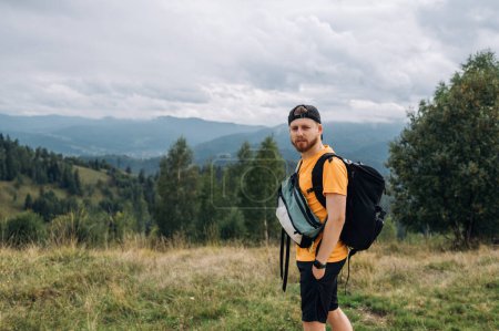 Téléchargez les photos : Portrait d'un beau jeune touriste avec une barbe en vêtements décontractés debout dans les montagnes sur le fond d'un beau paysage et regardant la caméra - en image libre de droit