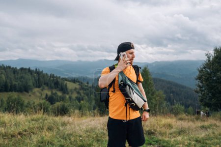 Téléchargez les photos : Un beau jeune homme en vêtements décontractés se tient dans les montagnes avec un smartphone et regarde loin avec un visage sérieux sur un fond de beaux paysages. - en image libre de droit