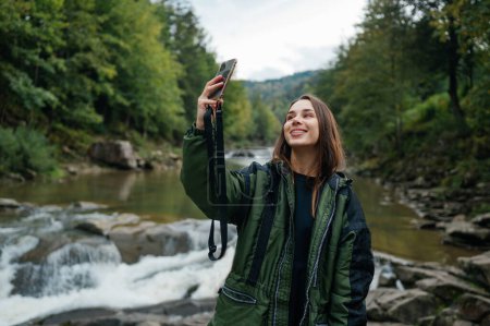 Photo for Happy female hiker taking selfie on smartphone against mountain river background while hiking and smiling. Mountain tourism. - Royalty Free Image