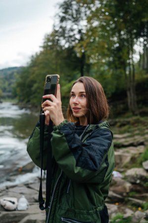 Photo for Portrait of positive woman hiker taking photo on smartphone camera with smile on face and smiling against background of mountain rapid river - Royalty Free Image