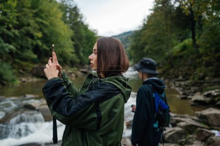 Photo for A couple of tourists are standing near a mountain river on the background of a forest, a woman is taking a photo on a smartphone camera on the background of a guy. Young tourists on a mountain hike. - Royalty Free Image