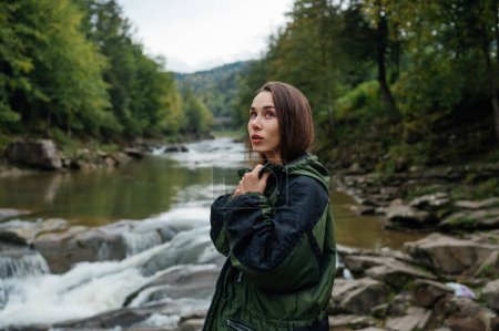 Photo for A beautiful female hiker on a hike stands on the background of a mountain river and forest and looks away with a serious face. - Royalty Free Image