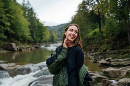 Téléchargez les photos : Une touriste féminine positive en vêtements décontractés se tient dans les montagnes sur la rive de la rivière avec un sourire sur le visage et regarde sur le côté sur le fond de belles vues sur la montagne. - en image libre de droit