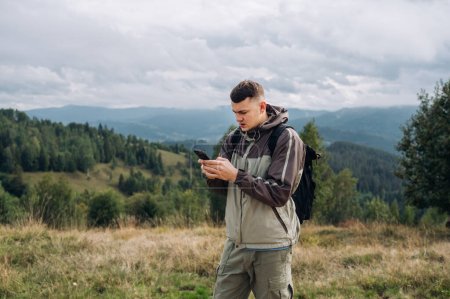 Téléchargez les photos : Un touriste masculin sérieux lors d'une randonnée se tient dans les montagnes sur le fond d'un beau paysage et utilise un smartphone avec un visage sérieux. - en image libre de droit