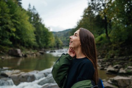 Photo for Happy woman hiker in a jacket in the mountains on a hike looks away with a surprised face and smiles against the background of a rocky mountain river and forest. - Royalty Free Image