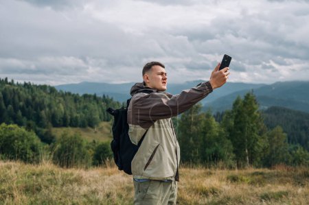 Photo for A serious male tourist in casual clothes stands with a smartphone in his hand in the mountains on the background of beautiful views and searches the network. - Royalty Free Image