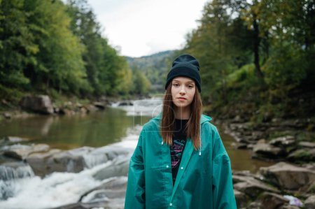 Photo for Attractive woman in raincoat and hat standing in mountains near river and rocks and posing for camera with serious face. - Royalty Free Image